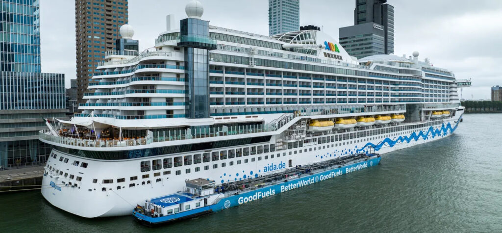 AIDAprima cruise ship docked in Rotterdam being bunkered with biofuel, with city skyscrapers in the background.